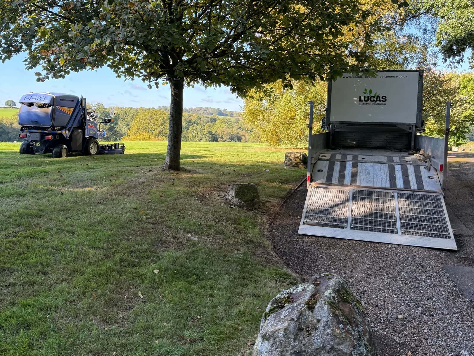 A white Lucas box trailer with its ramp down beside a ride-on mower on parkland grass