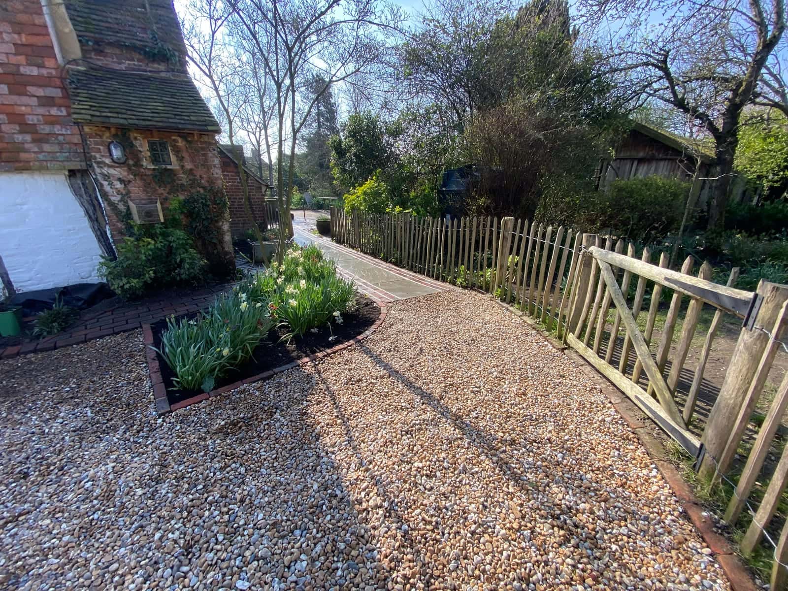A chestnut paling fence and gate beside a gravel path and spring planting outside an old cottage