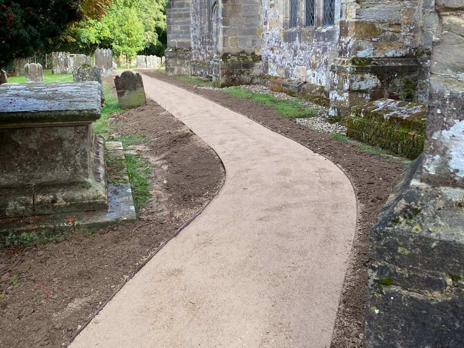 A finished buff-coloured path curving along the stone wall of a church, past a chest tomb