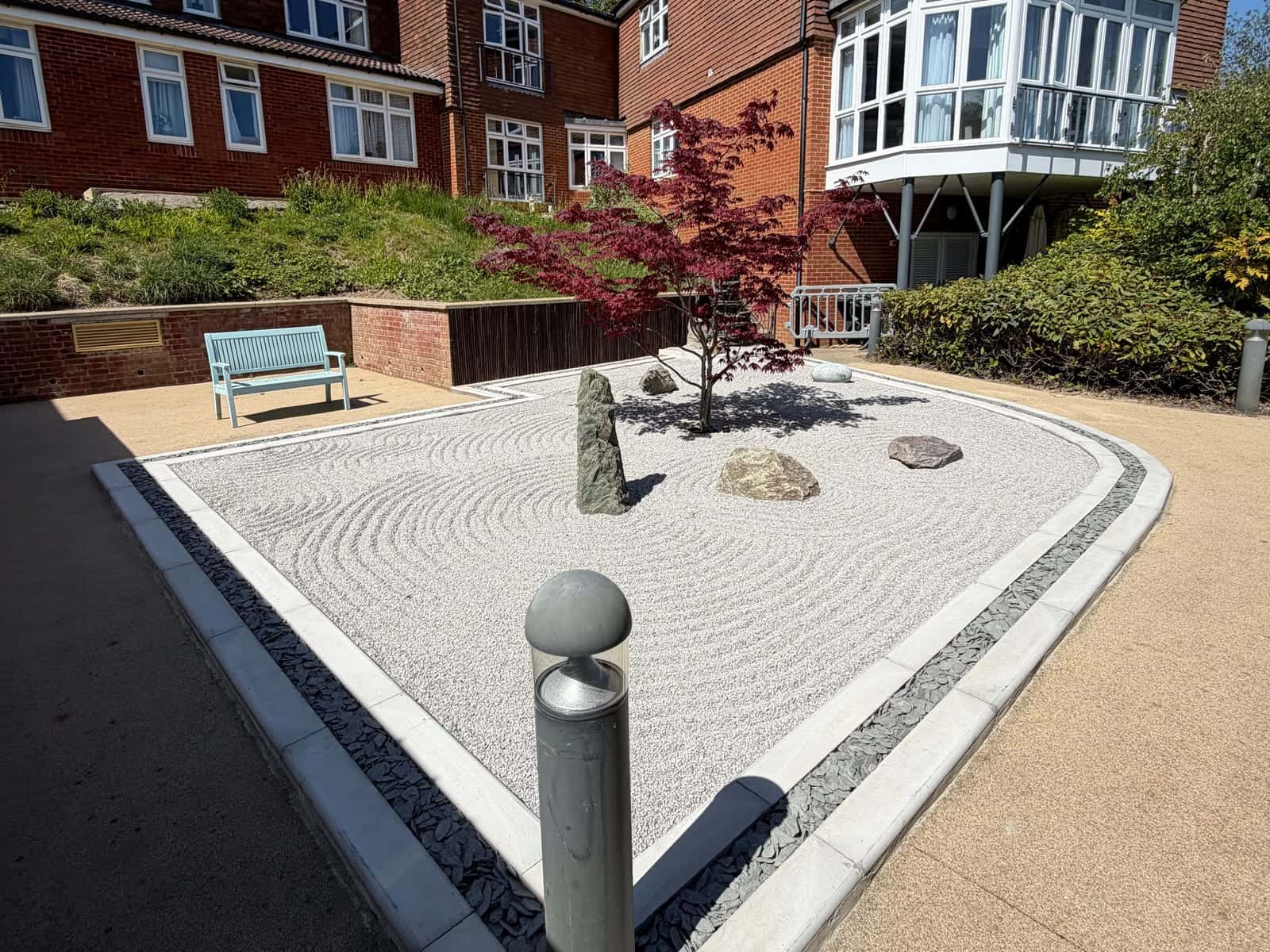 A raked gravel garden with a red acer, standing stones and a slate border, in a courtyard between brick buildings