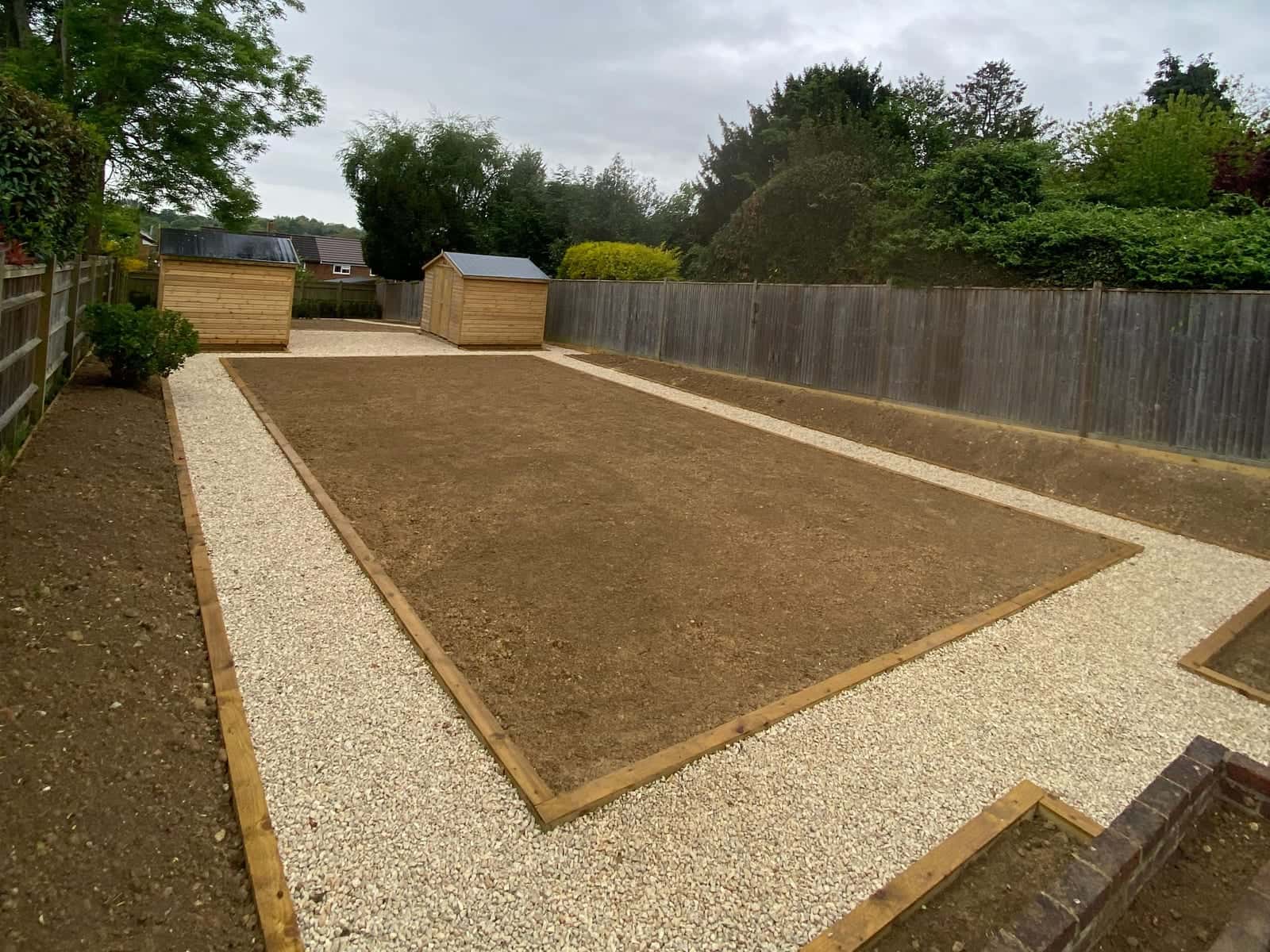 Gravel paths with timber edging around a large rectangle of levelled soil ready for turf, with two timber sheds and a fence beyond