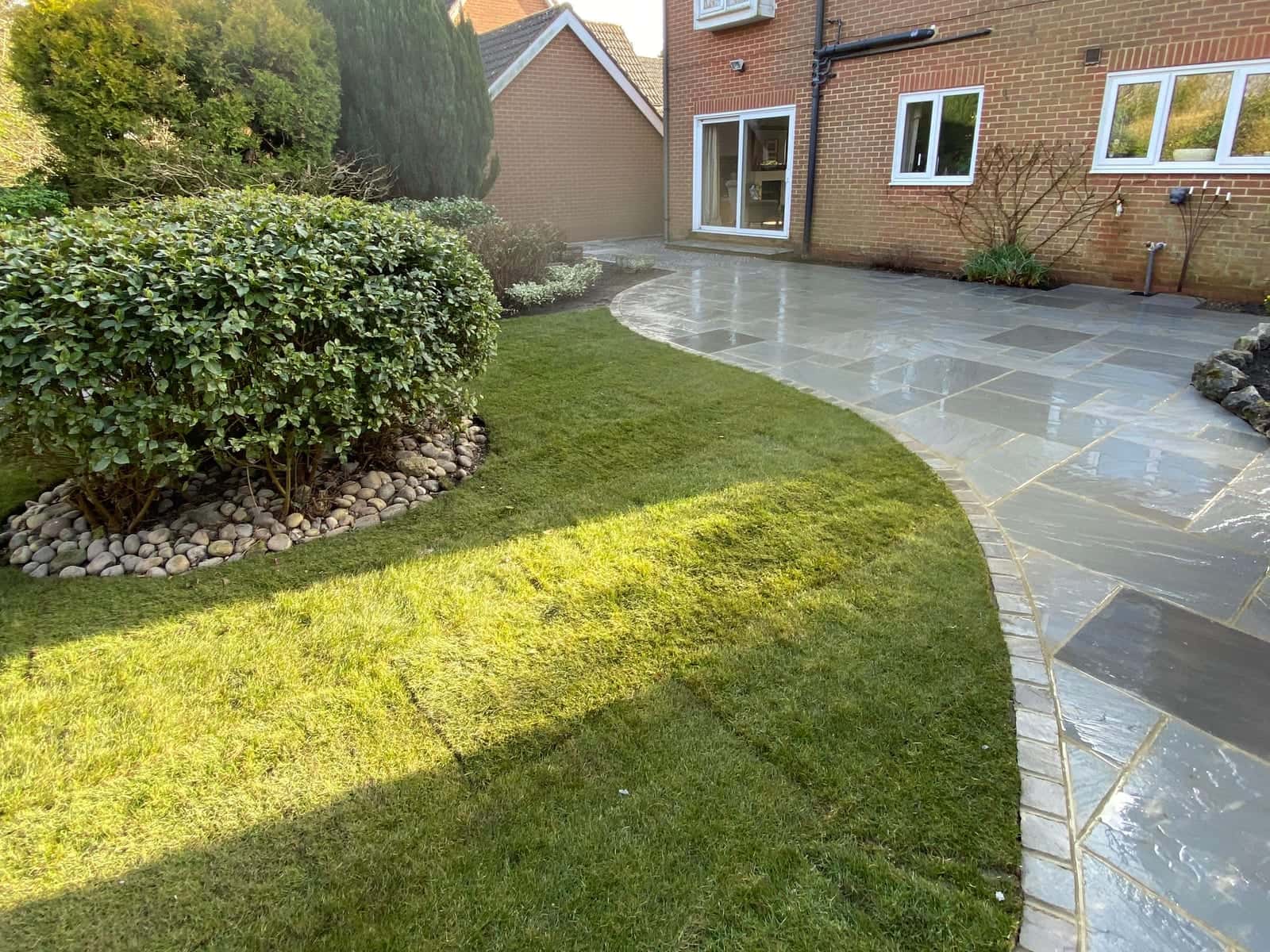 A curving grey sandstone path beside a new lawn and a clipped shrub, leading to the back of a brick house