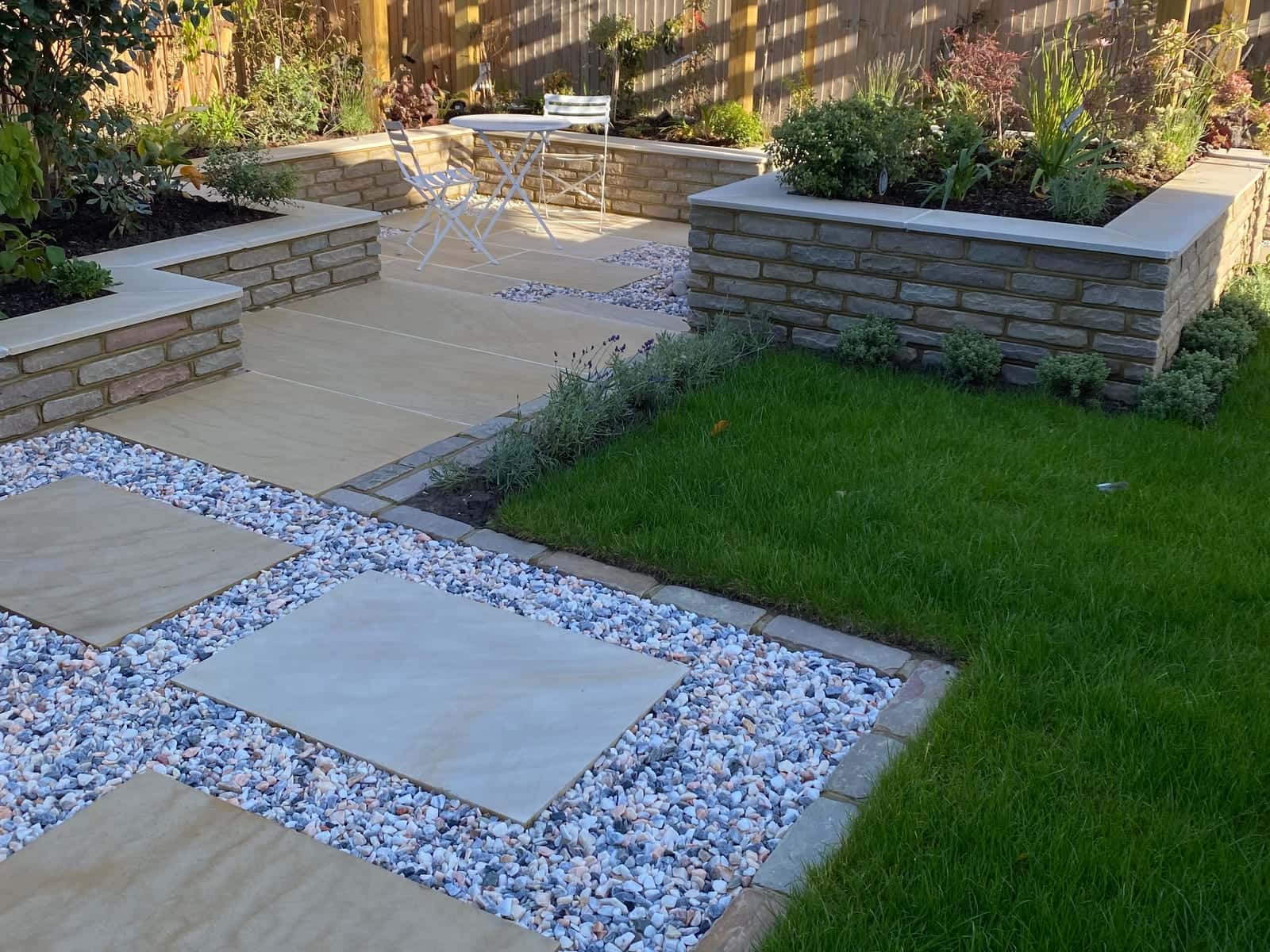 Sandstone paving and large stepping stones set in pale gravel, with raised stone-walled planting beds, a timber pergola and new turf, in a garden backed by modern brick houses