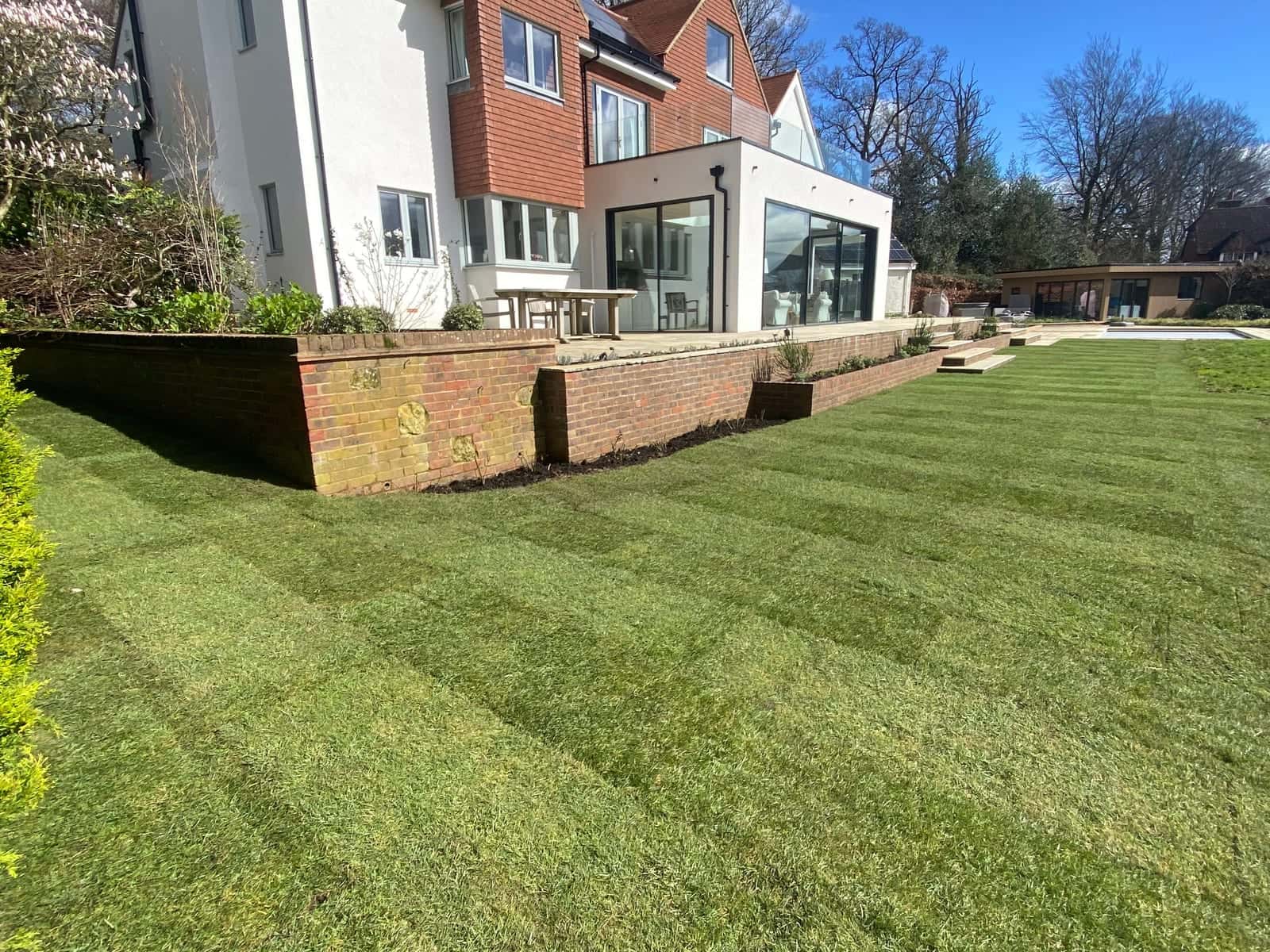 A new striped lawn below brick terrace walls at the back of a large rendered and tile-hung house