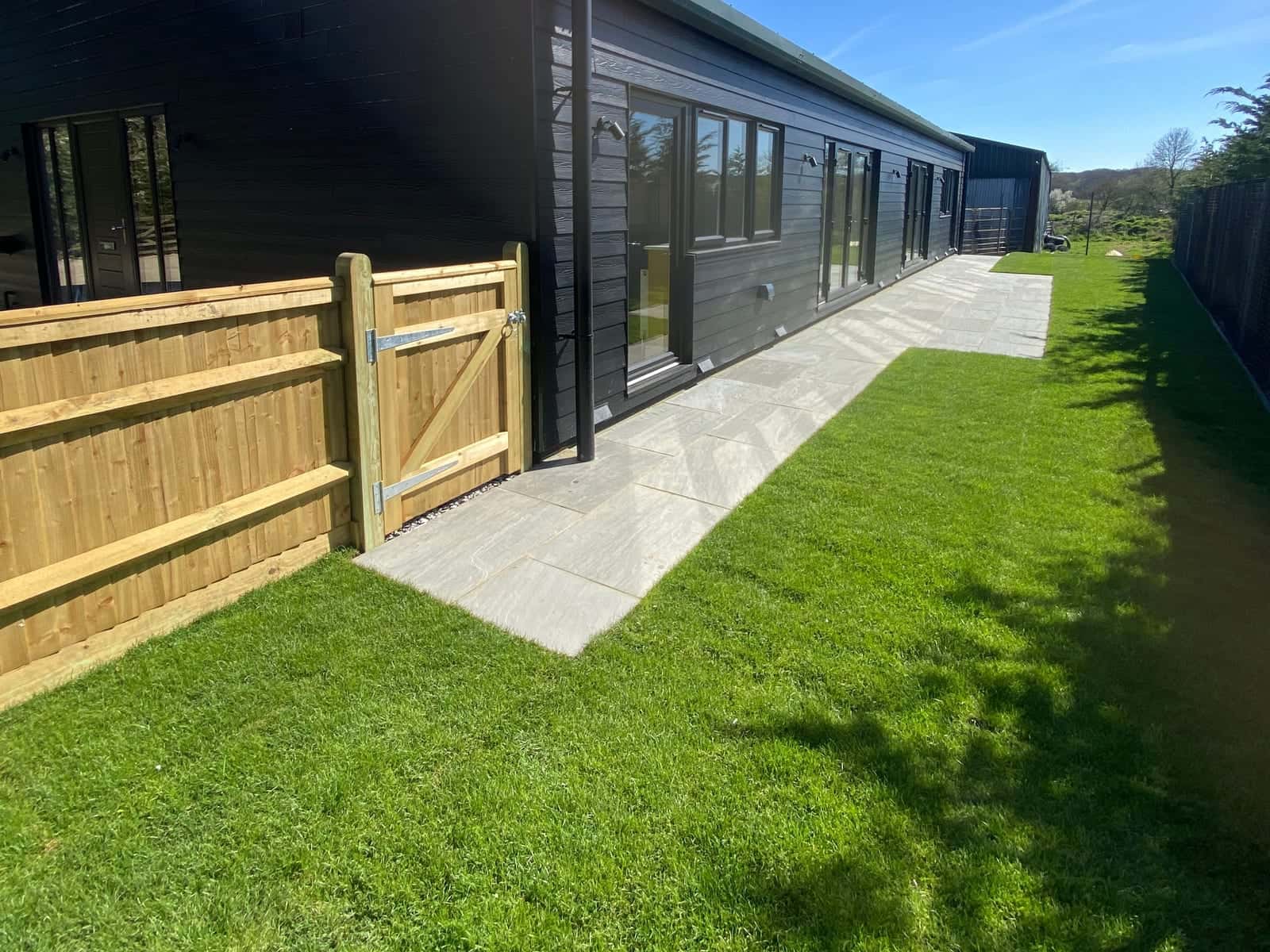 A new lawn and paved path along a dark timber-clad building, with a timber gate and fence in the foreground