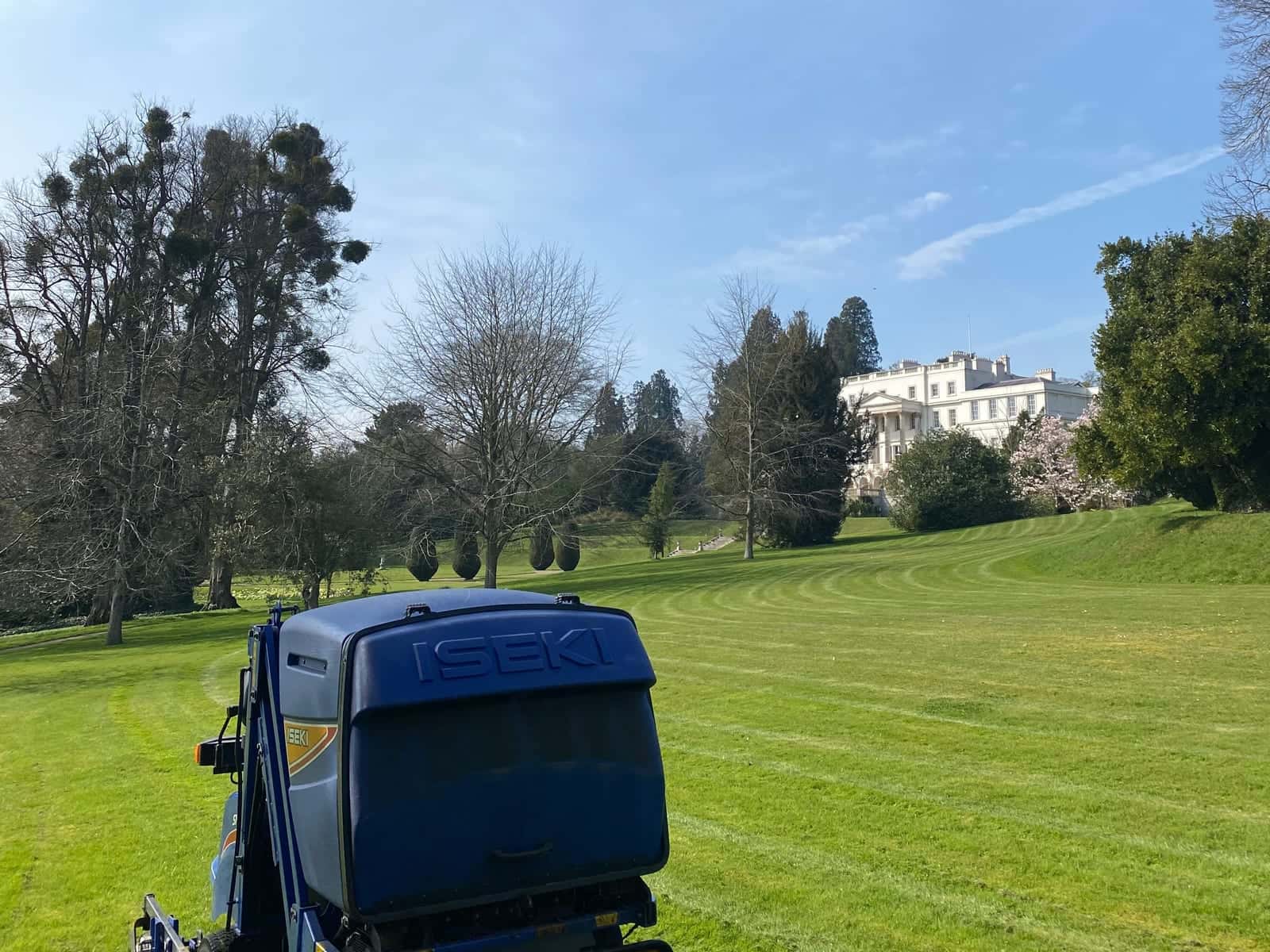A ride-on mower on striped parkland grass, with specimen trees and a large white house in the distance