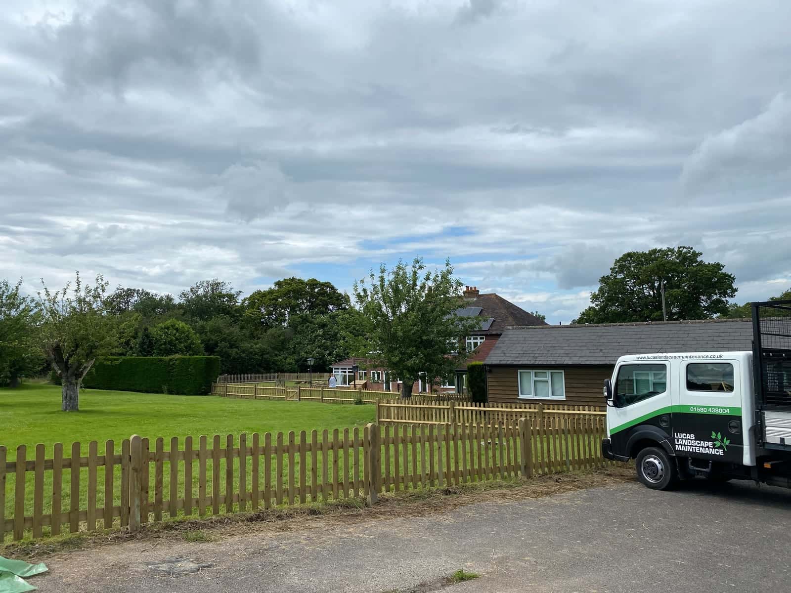 Timber picket fencing around a large lawn, with a house and trees beyond and a Lucas Landscape Maintenance truck parked on the drive