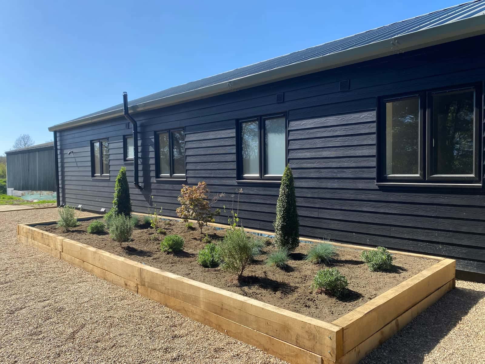 A raised timber sleeper bed of young shrubs and grasses beside a dark timber-clad building, on gravel