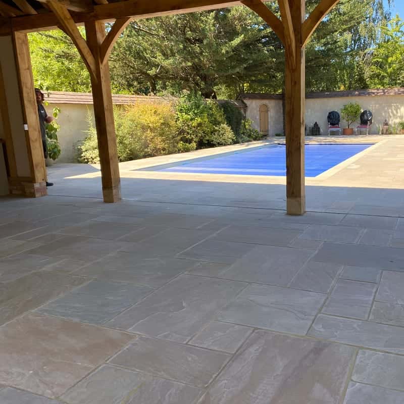 Sandstone paving under an oak-framed pavilion beside a covered swimming pool, with potted plants along a garden wall