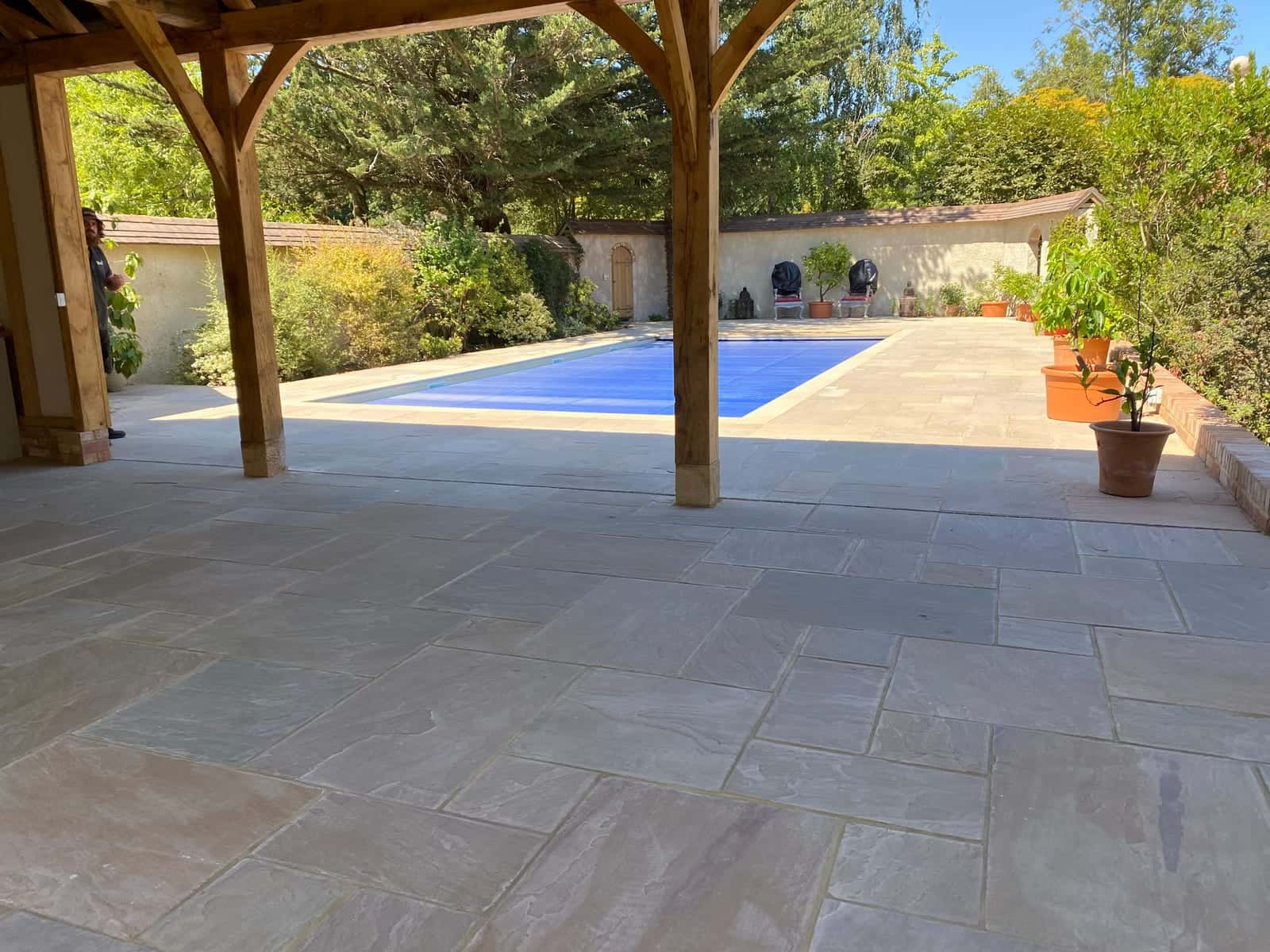 Sandstone paving under an oak-framed pavilion beside a covered swimming pool, with potted plants along a garden wall
