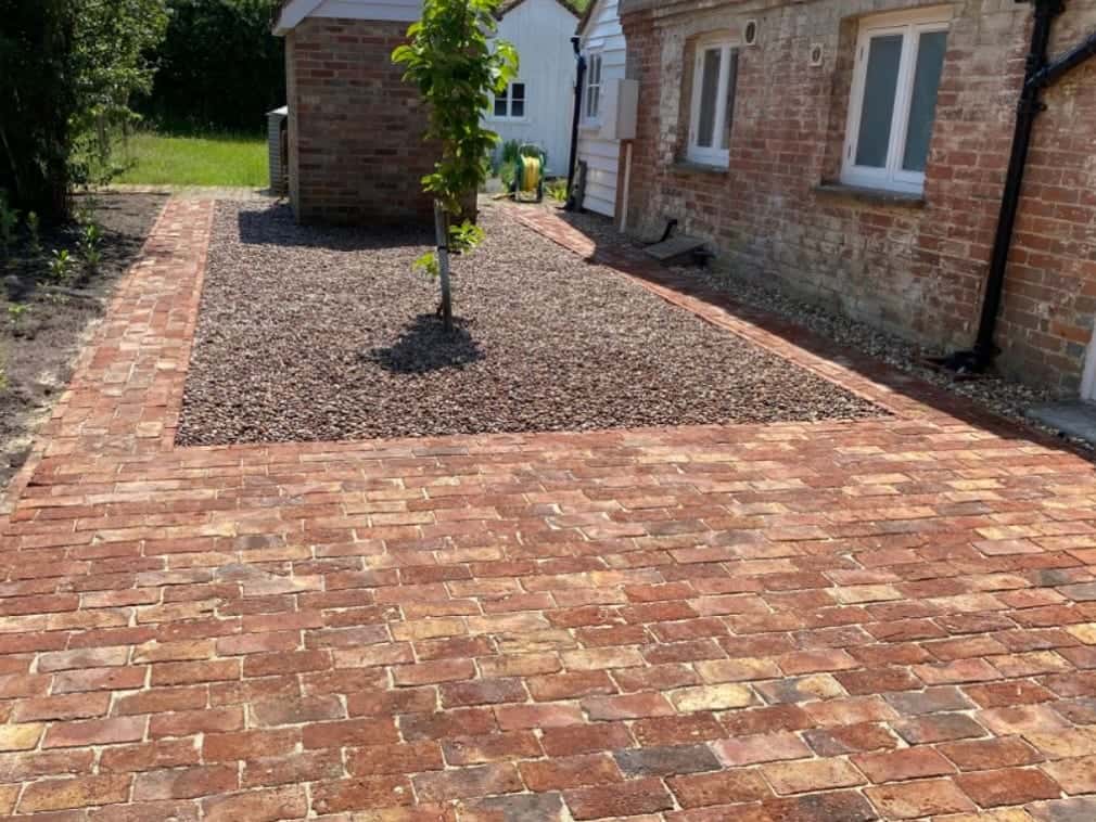 Reclaimed brick paving around a gravel bed planted with a young tree, beside an old brick house and a weatherboarded outbuilding