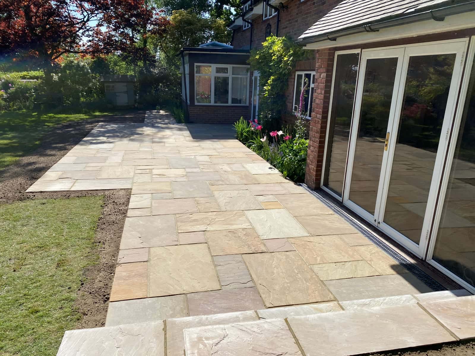A newly laid sandstone patio running along bifold doors at the back of a brick house, with lawn to one side