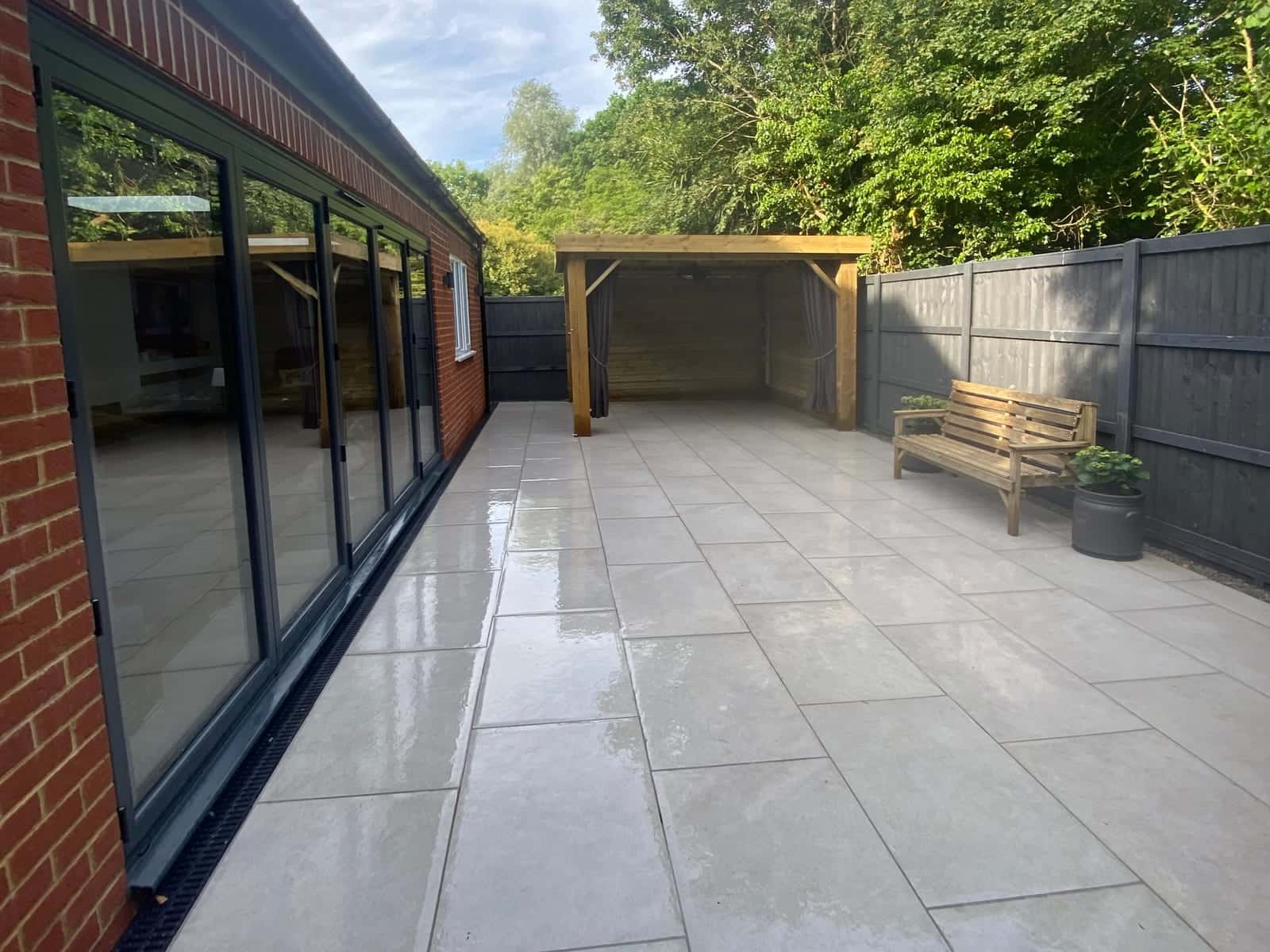 Pale paving along the glazed side of a brick building, with a timber pergola and a bench at the far end
