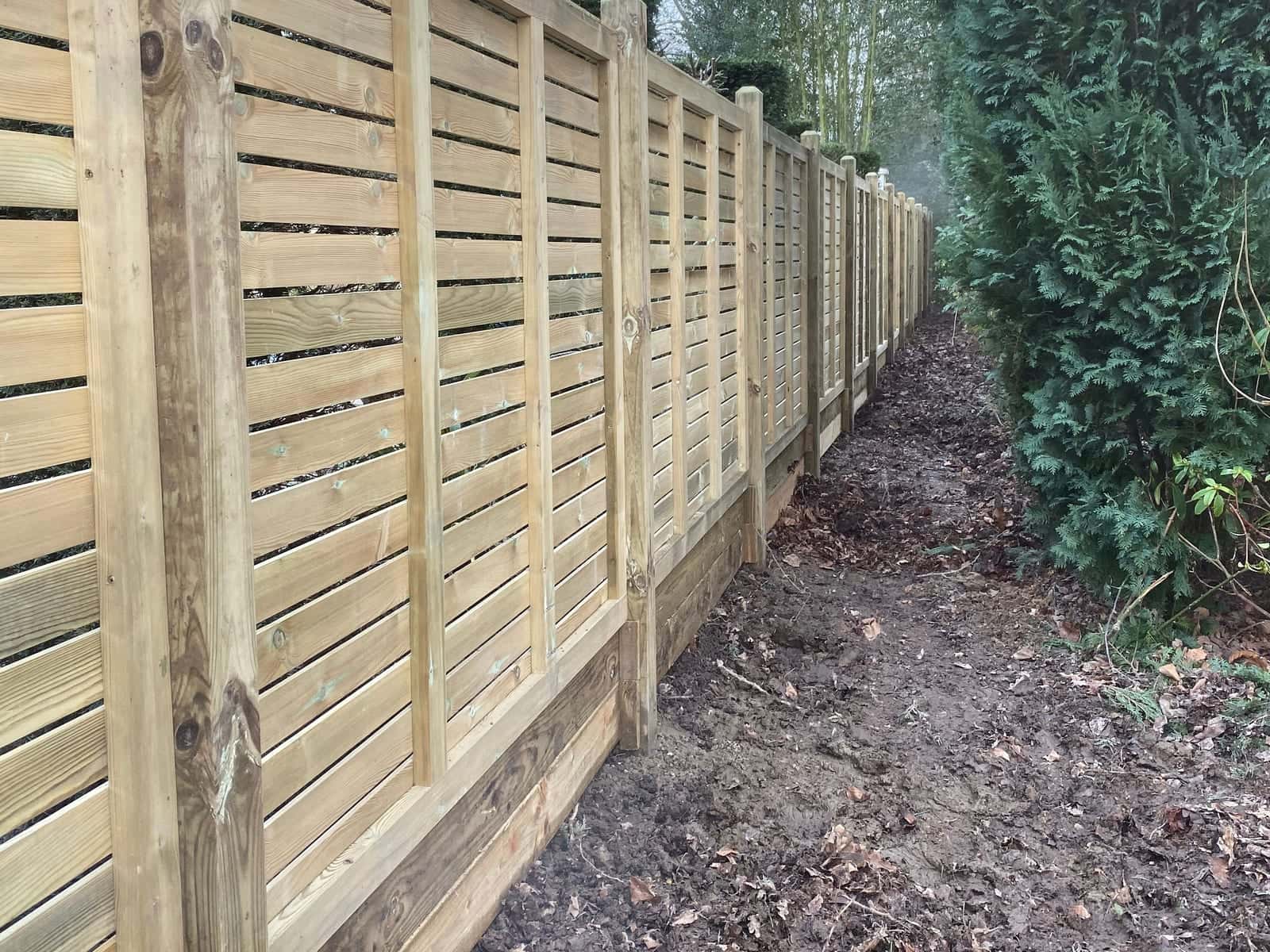 A long run of new slatted timber fencing on square posts along a woodland boundary