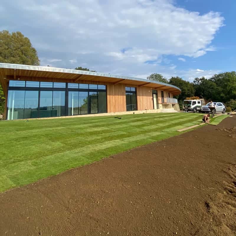 Fresh turf laid across a new lawn in front of a long timber-clad house with a curved roof, with bare prepared ground still to be turfed in the foreground