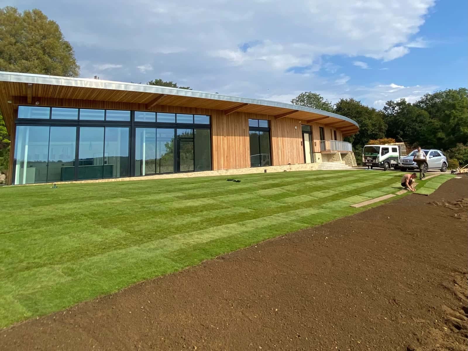 Fresh turf laid across a new lawn in front of a long timber-clad house with a curved roof, with bare prepared ground still to be turfed in the foreground
