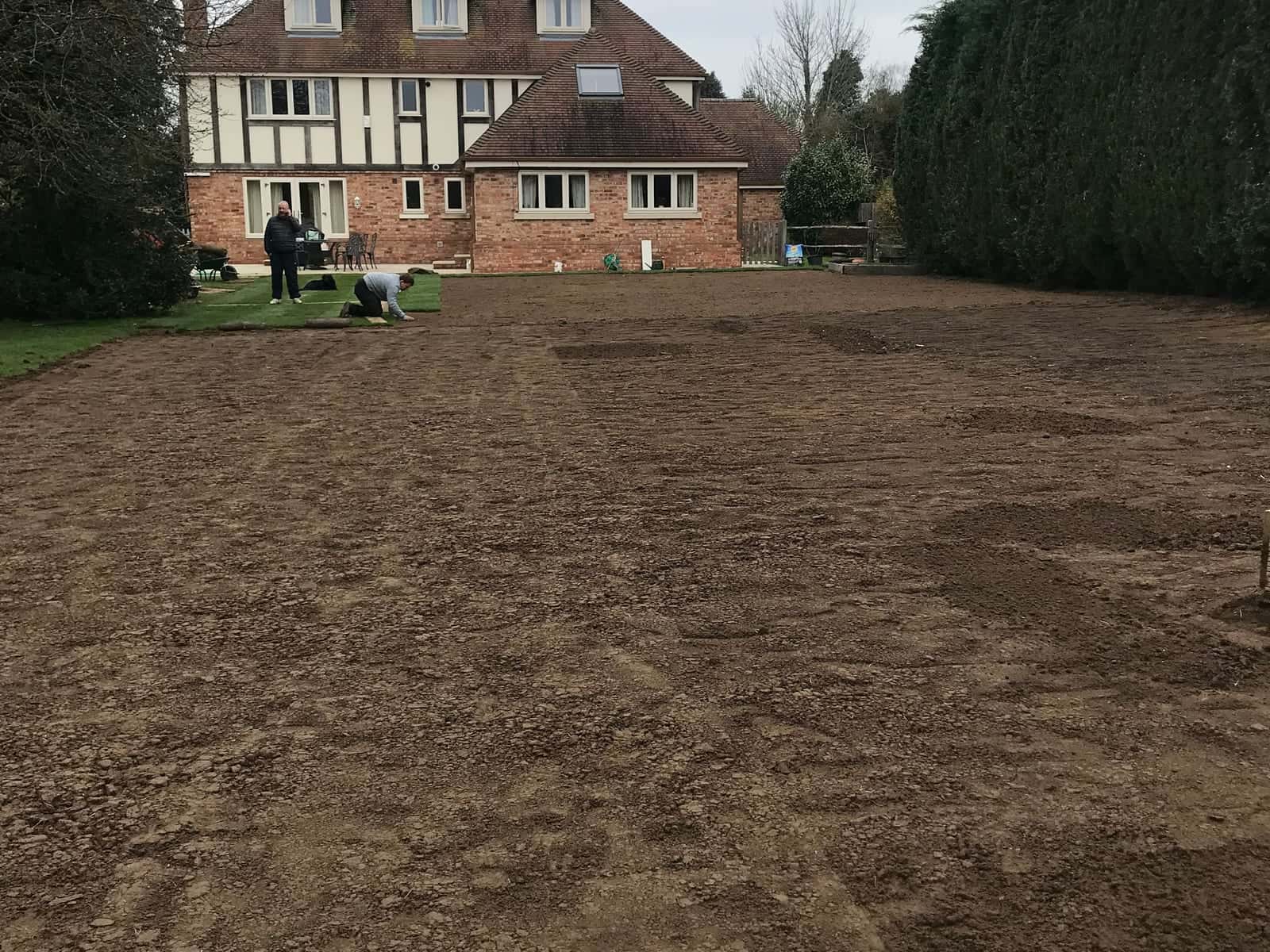A large rear garden raked level for turf, with two people working near a mock-Tudor house