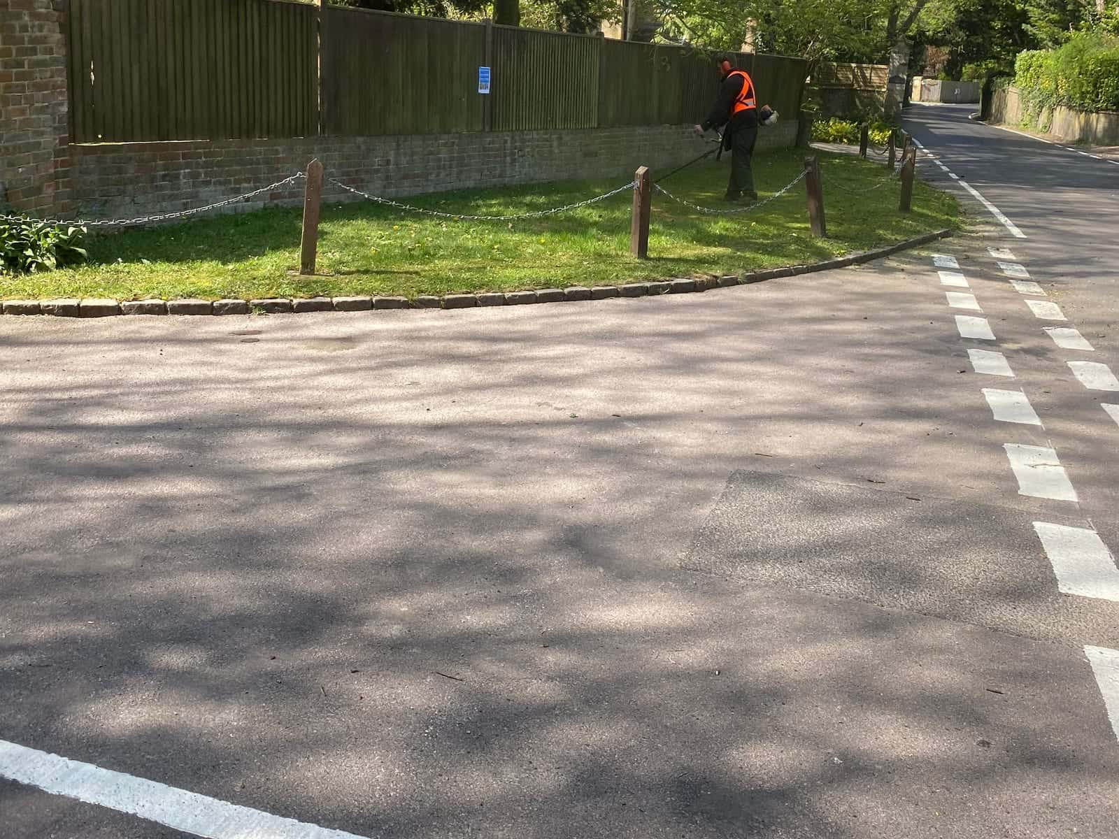 An operator in high-visibility clothing strimming a grass verge beside a road and a brick and timber boundary