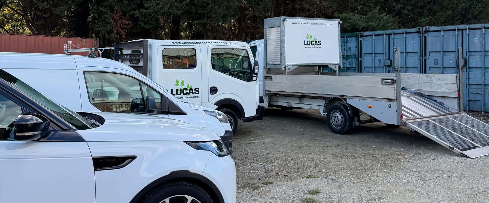 Three white Lucas Landscape Maintenance vehicles, a 4x4, a crew-cab tipper and a box van with its ramp down, on gravel hardstanding