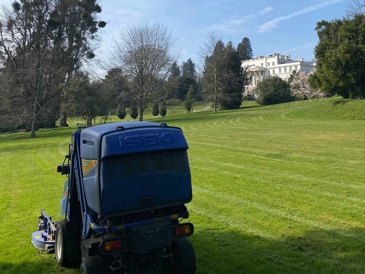 A blue Iseki ride-on mower on a freshly striped parkland lawn, with mature trees and a white country house beyond under a spring sky