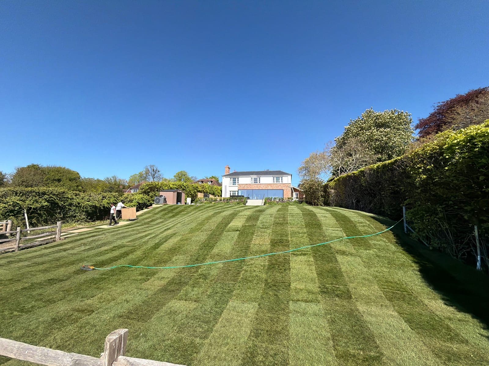 Freshly laid turf striped across a large, gently sloping garden, with a house at the far end and a hose across the lawn