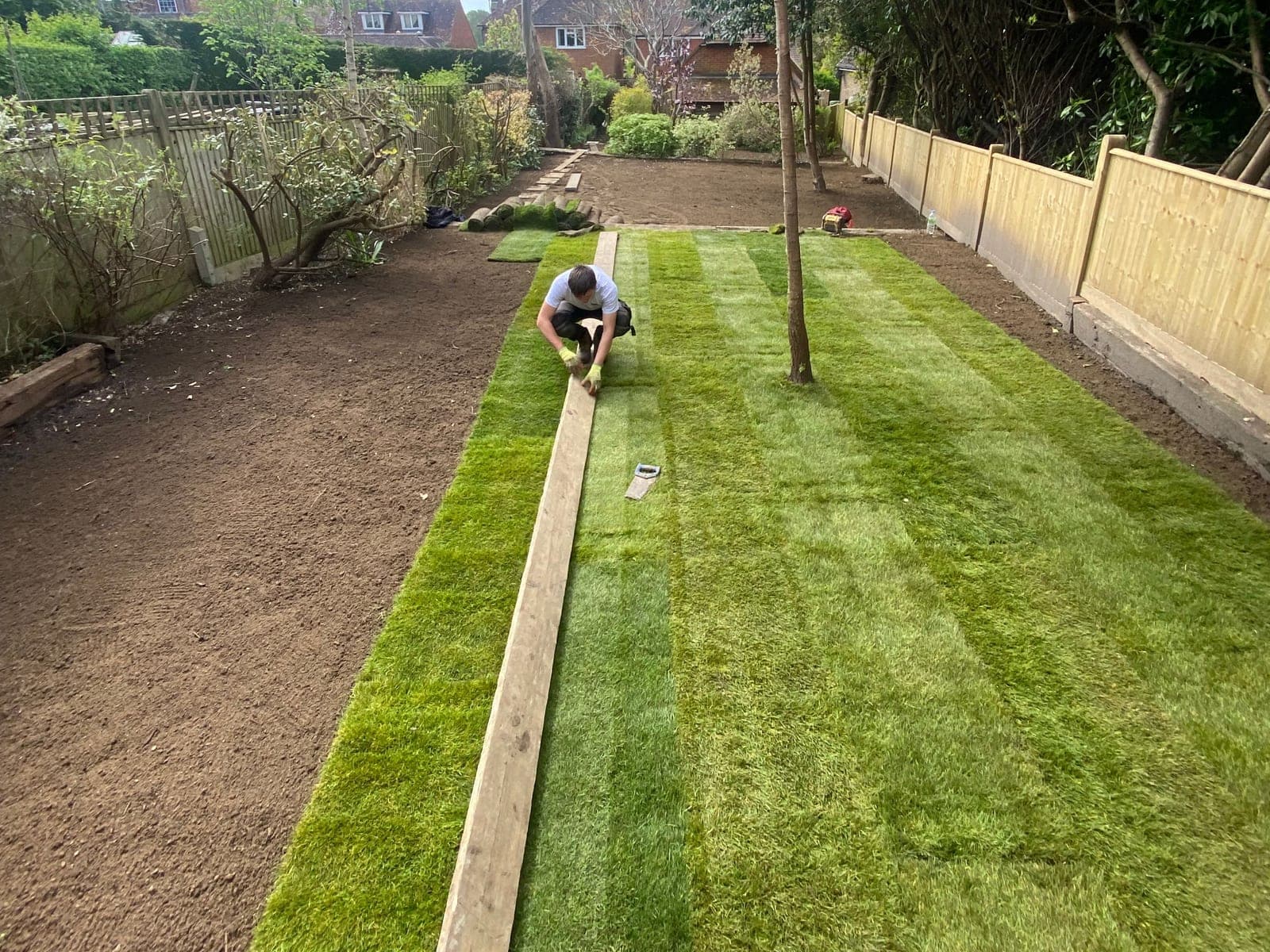A landscaper kneeling to lay rolls of turf down a long garden beside a timber edging board