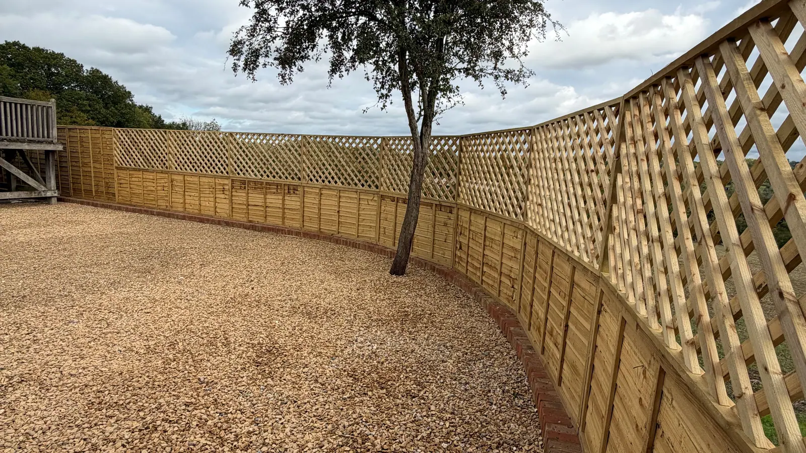 A new curving fence with trellis panels along the top, enclosing a gravel garden around a single tree