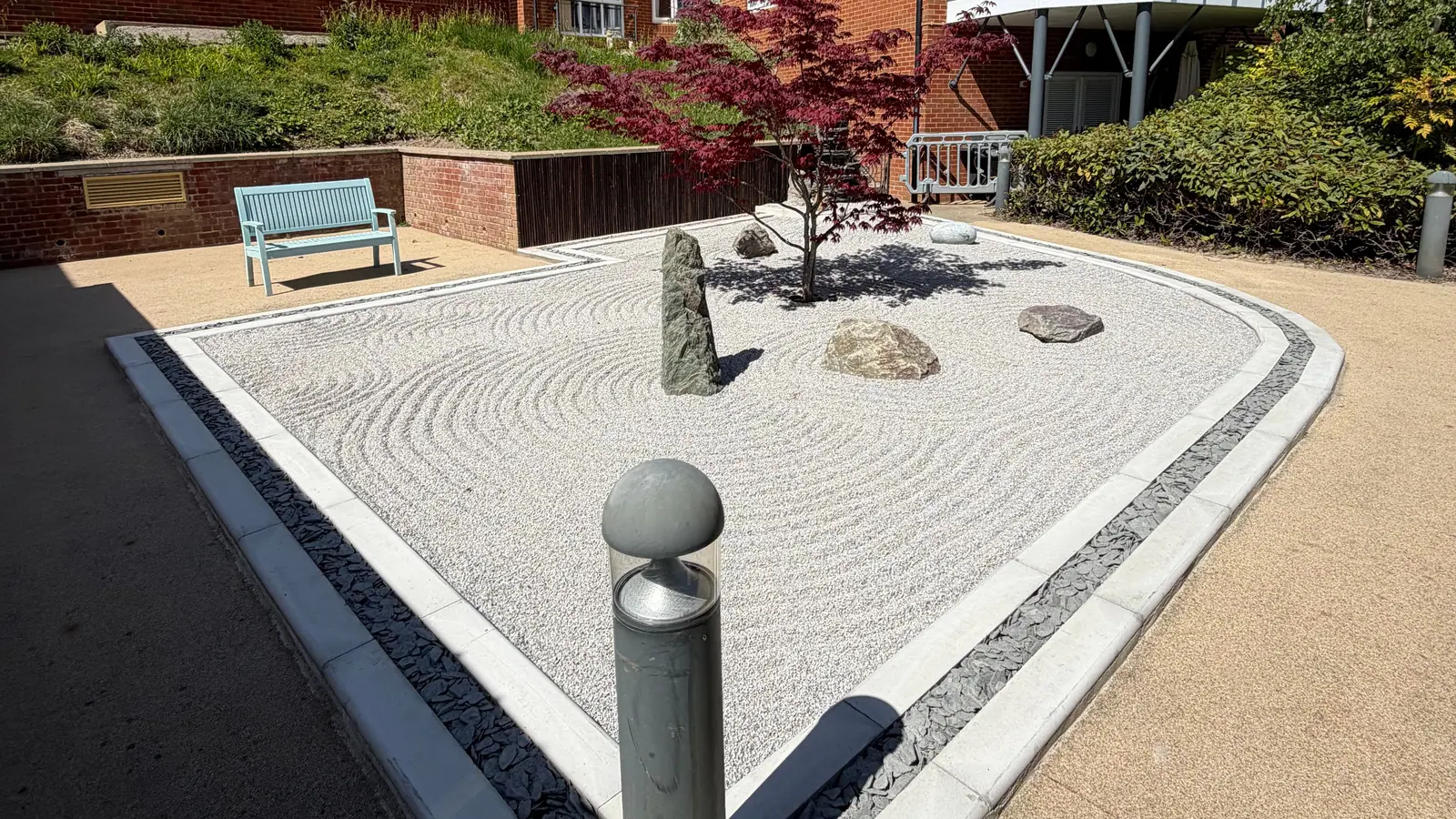 A raked gravel garden with a red acer, standing stones and a slate border, in a courtyard between brick buildings