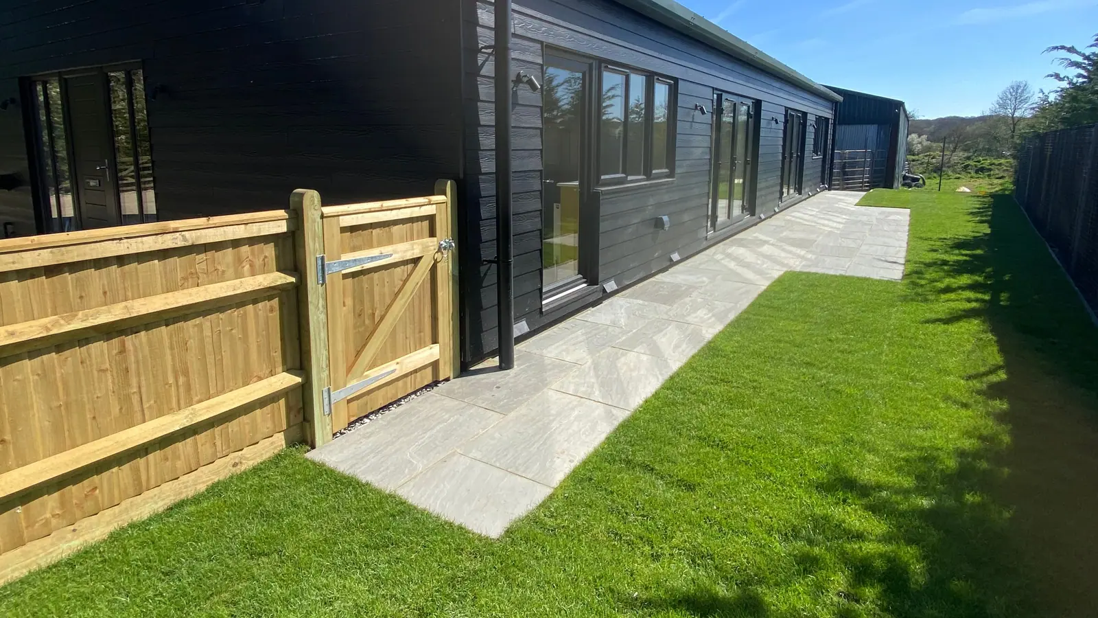 A new lawn and paved path along a dark timber-clad building, with a timber gate and fence in the foreground
