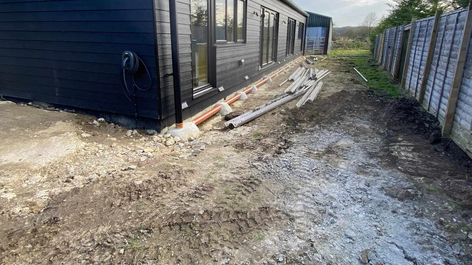 Bare, rutted ground and building offcuts along the side of a dark timber-clad building, before work began